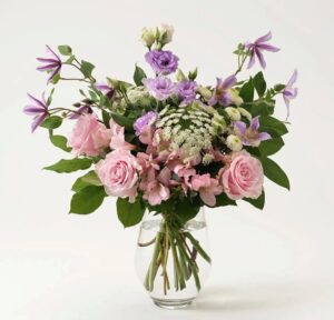Rosa rosor, ljuslila clematis och prärieklockor tillsammans med dekorationsgrönt. En härlig försommarbukett! Skicka med bud via Interflora!